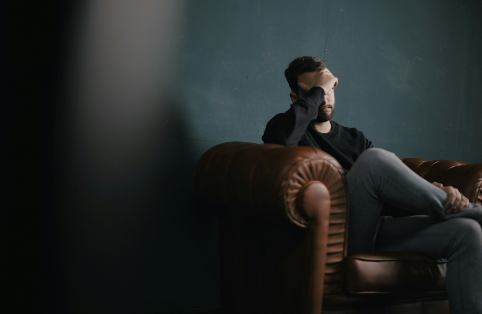 crisis services man sitting on couch in dark room with face resting in hand.